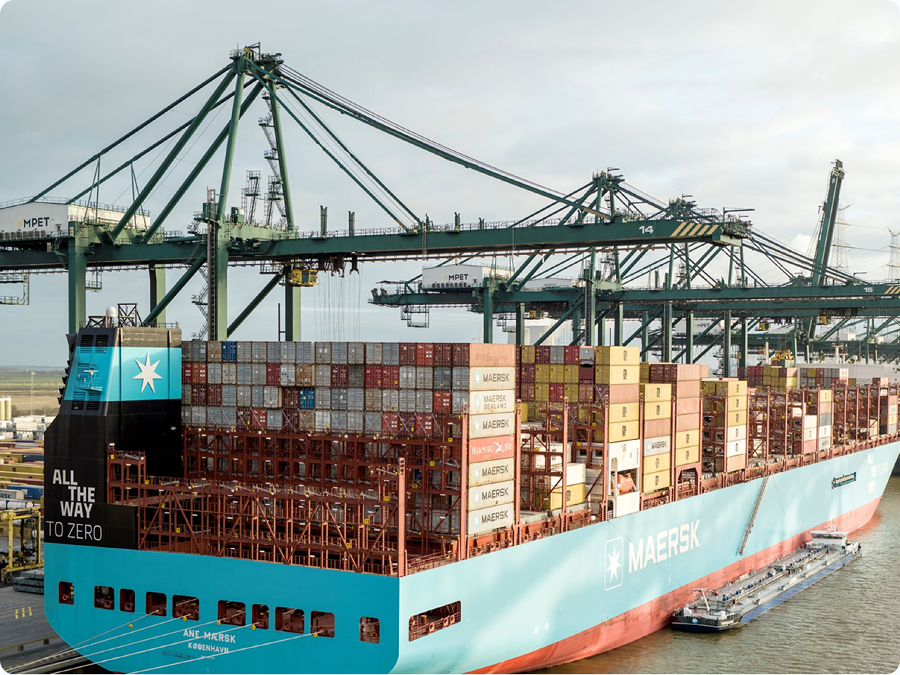 Een groot lichtblauw Maersk-containerschip, de Ane Mærsk, ligt aangemeerd bij de MPET-terminal. Op de achtergrond staan meerdere groene containerkranen. Het schip is zwaar beladen met diverse gekleurde containers en op de achtersteven staat de tekst 'All the way to zero'.