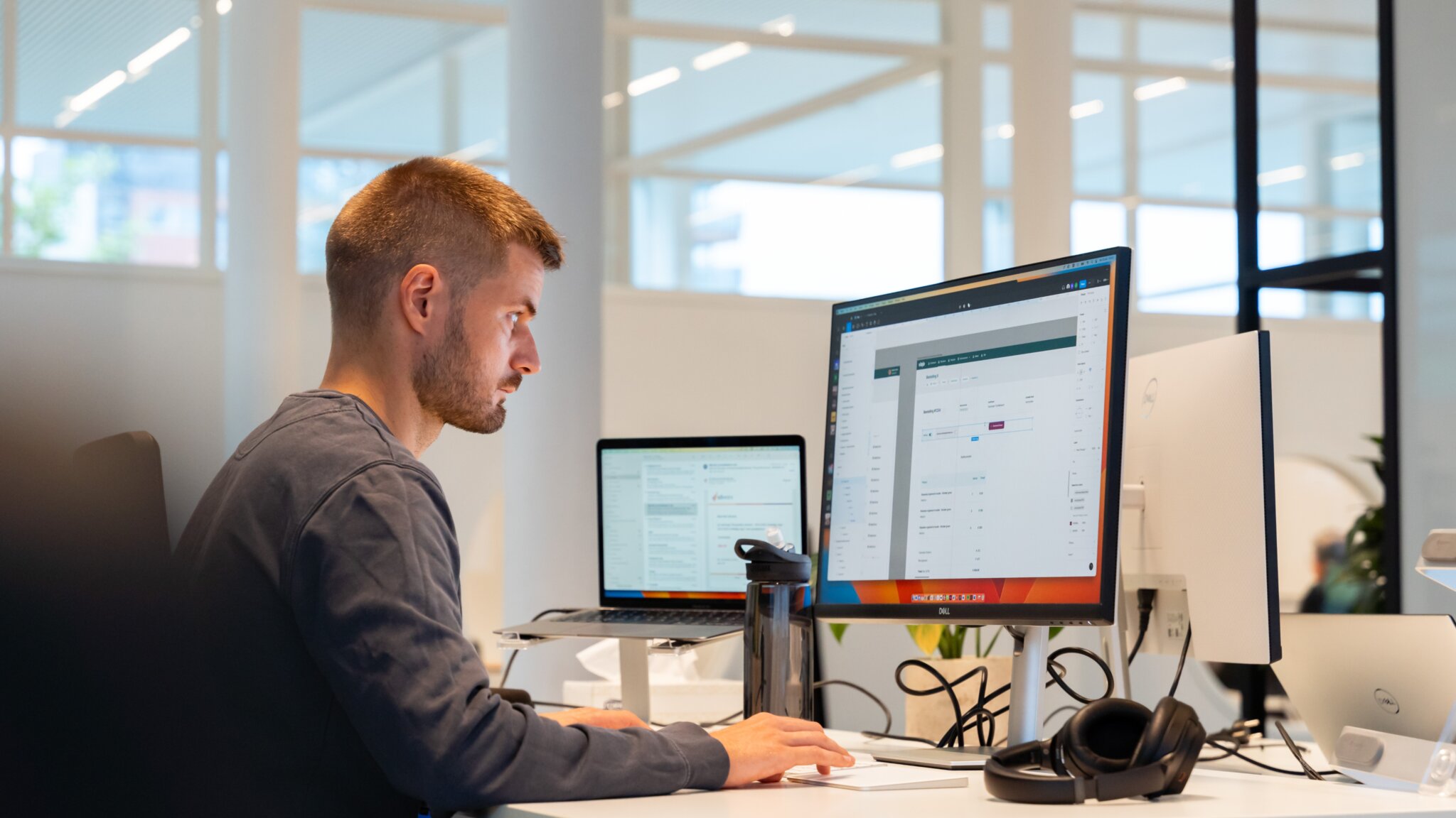 UX'er bert werkt aan een user interface ontwerp op een groot computerscherm in een lichte kantooromgeving met een laptop en koptelefoon op het bureau.